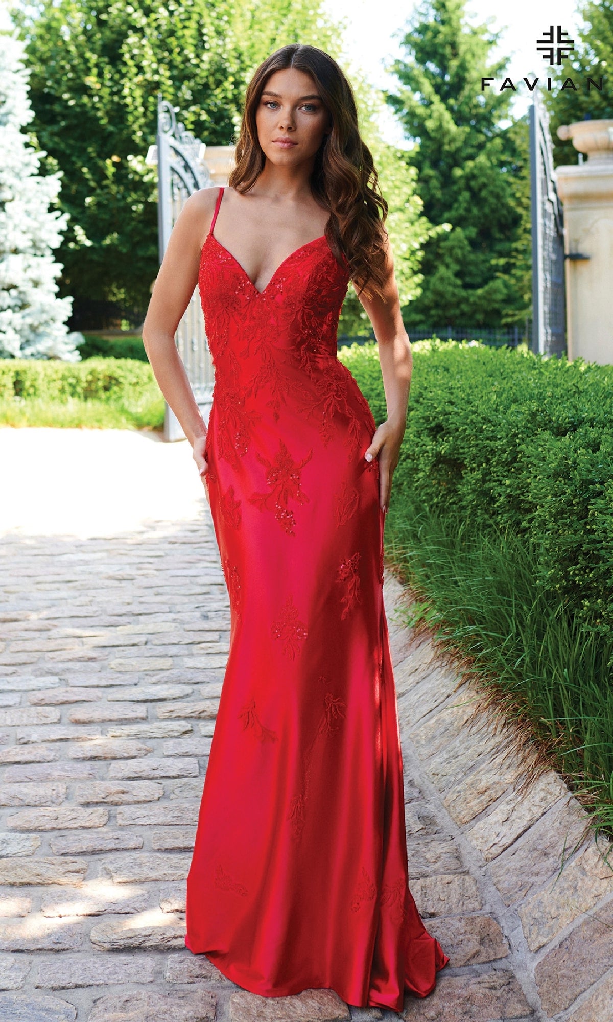 A woman stands outdoors on a stone path wearing the Faviana 11245 Strappy Open-Back Long Prom Dress in red, featuring thin straps and floral embroidery. Lush greenery and an open gate are visible in the background.